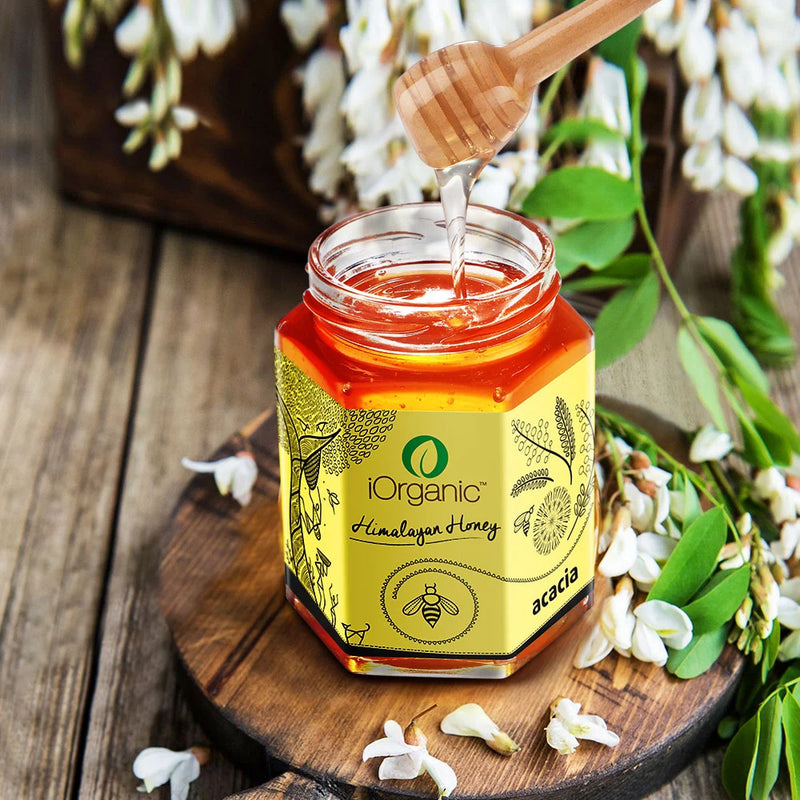 Freshly extracted iOrganic Acacia Honey being poured from a honey dipper, surrounded by Acacia flowers.